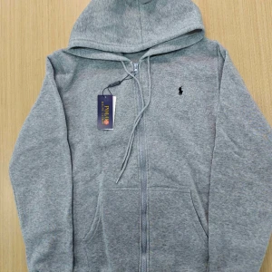 Grå hoodie från Polo Ralph Lauren - Säljer en klassisk grå hoodie från Polo Ralph Lauren med dragkedja framtill och justerbar huva. Tröjan har en liten broderad logga på bröstet och två fickor framtill. Perfekt för en avslappnad stil.