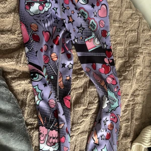 Lila leggings med pop art-mönster från Stronger - Färgglada leggings i lila med pop art-inspirerat mönster med hjärtan, stjärnor och tecknade detaljer. Hög midja och stretchigt material som passar perfekt till träning eller en färgstark vardagslook.fint skick 