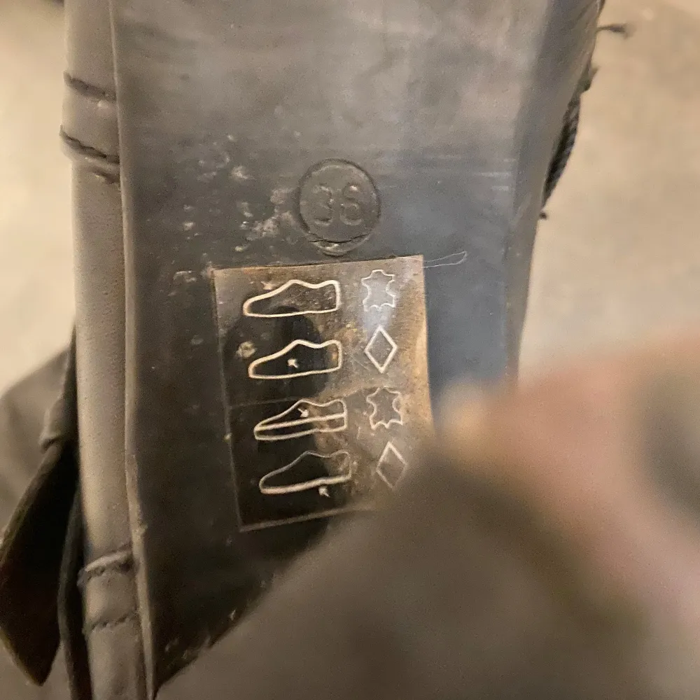 Säljer ett par svarta höga stövlar med dekorativa spännen och dragkedja på sidan. Skorna har klack och är tillverkade i äkta skinn. Knappt använda de är i storlek 36. Kengät.