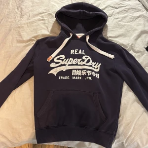 Mörkblå hoodie från Superdry - Säljer en mörkblå hoodie från Superdry med vit logga och text framtill. Tröjan har huva med vita dragsnören och en stor magficka. Perfekt för dig som gillar en avslappnad och sportig stil.