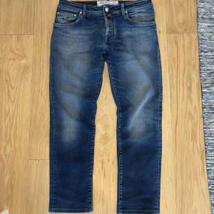 Jacob Cohën Jeans Size: 33 - Säljer ett par klassiska blå jeans från Jacob Cohën med snygg tvätt och raka ben. Jeansen har fem fickor, knappgylf och diskreta slitningar för en cool look. Perfekta för dig som gillar italiensk design och exklusiva material.