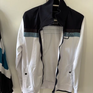 Vit och svart track jacket från Jack & Jones - Säljer en snygg track jacket från Jack & Jones i vitt och svart med blå och bruna detaljer. Jackan har dragkedja, hög krage och två fickor framtill. Stort tryck med siffran 89 på ryggen. Perfekt för en sportig stil.
