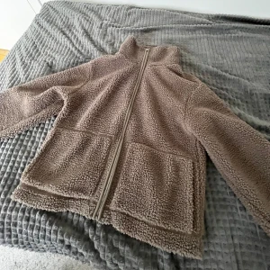 Beige teddyjacka med dragkedja - Mysig beige teddyjacka med hög krage och hel dragkedja framtill. Jackan har två stora fickor på framsidan och är långärmad. Perfekt för kyligare dagar.