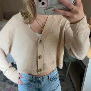 Beige croppad kofta med knappar - Säljer en beige croppad kofta med stora knappar framtill och djup v-ringning. Tröjan är stickad och har långa ärmar samt ribbade muddar. Perfekt att styla med jeans eller kjol.