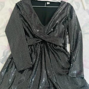 Glittrig silverklänning med v-ringning från H&M - Säljer en glittrig silverfärgad klänning från H&M med djup v-ringning och långa ärmar. Klänningen har omlott-detalj framtill och markerad midja. Perfekt för festliga tillfällen!