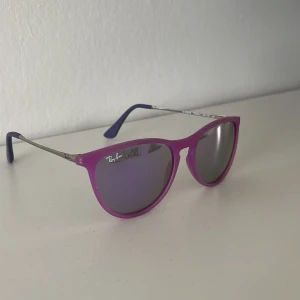 Lila solglasögon från Ray-Ban - Säljer ett par snygga solglasögon från Ray-Ban med lila bågar och mörka glas. Bågarna har en klassisk rundad form och tunna metallskalmar. Perfekt accessoar för att lyfta din stil. Använda ett fåtal gånger. Barn storlek!!