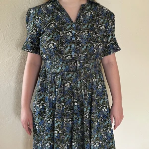 Vintage blommig klänning med bälte och knappar - Retroinspirerad klänning med blått och grönr blommönster, korta puffärmar och v-ringning. Knappar framtill och matchande bälte i midjan. Perfekt för dig som gillar vintage vibes och vill sticka ut med en färgglad look. Väldigt bra skick. 