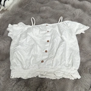 Vit offshoulder topp med knappar - Säljer en söt vit offshoulder topp med smala axelband och korta ärmar. Toppen har knappar framtill, smockad nederkant och volangdetalj längst ner. Perfekt för varma dagar och enkel att matcha.