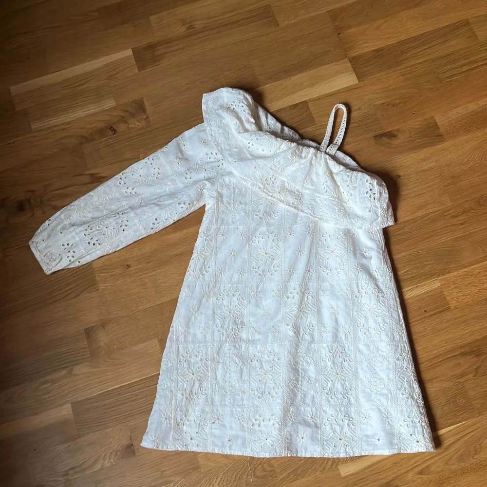 Superfin vit klänning från Zara med broderade blommor och mönster. Klänningen har en ärm och en axelbandslös sida med volangdetalj upptill. Perfekt för sommarens tillfällen och har en oneshoulder-design.. Mekot.