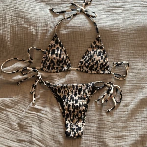 Leopardmönstrad bikini med knyt, oanvänd  - Säljer en snygg bikini med leopardmönster i svart, beige och vitt. Bikinin har trekantsöverdel med knyt i nacken och ryggen samt matchande underdel med knyt i sidorna. Materialet är smockat och stretchigt för en bekväm passform..