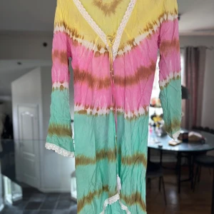 Färgglad långklänning från Hot Lava  - Säljer en långklänning i bohemisk stil från hot lava  med batikmönster i gult, rosa och turkost. Klänningen har v-ringning med spetsdetaljer och knyt framtill. Perfekt för sommarens alla tillfällen!