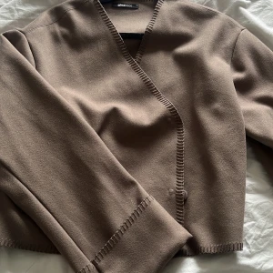 Brun/beige omlottjacka från Ginatricot - Säljer en brun/ beige omlottjacka från Ginatricot med dekorativa synliga sömmar och knappar på insidan. Jackan har lång ärm och en croppad passform, perfekt för lager på lager. Enkel och stilren design som passar till mycket.
