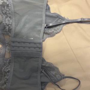 Blå  spetsig korsett-topp - Säljer en snygg blå korsett-topp med blommig spets och mesh-detaljer. Toppen har justerbara axelband, små rosetter framtill och hakknäppning bak. Perfekt för att lyfta din outfit med en romantisk touch.