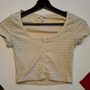 Beige croppad topp med knappar från H&M - Säljer en söt beige croppad topp från H&M med korta ärmar och dekorativa knappar som går att knäppa upp. Toppen har ett hålmönster över hela tyget och en rundad halsringning.