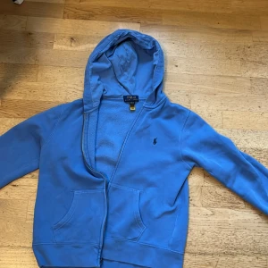 Blå hoodie från Polo Ralph Lauren - Säljer en blå hoodie från Polo Ralph Lauren med dragkedja och huva. Tröjan har två fickor framtill och den klassiska broderade loggan på bröstet. Perfekt för en avslappnad stil. Jag säljer denna p.g.a att den är för liten. Viktigt att veta den är storlek 10-12 år M och inte M vuxen storlek. Jag köpte denna från kidsbrandstore för 1199kr. Tröjan har bra skick och jag säljer denna för 349+frakt. Pris kan diskuteras 