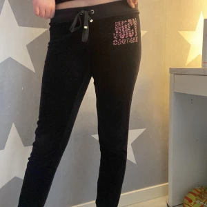 Svarta mjukisbyxor från Juicy Couture - Svarta mjukisbyxor från Juicy Couture med färgglatt leopardmönstrat logotryck på ena benet. Byxorna har dragsko i midjan och en ficka bak med knappar. Mjukt och skönt material, perfekta för en avslappnad stil.