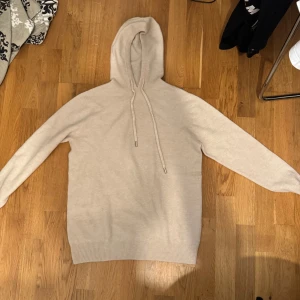 Beige stickad hoodie - Mysig beige hoodie med huva och dragsko. Tröjan är stickad och har långa ärmar samt breda ribbade muddar vid ärmslut och nederkant. Perfekt för mysiga dagar. Tröjan är aldrig använd. Koftan är i merinoull och i storlek M. 