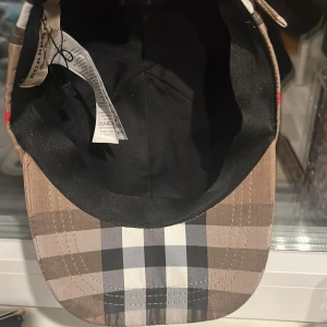 Rutig keps från Burberry - Säljer en klassisk rutig keps från Burberry med broderad logga baktill. Kepsen har justerbart spänne och är tillverkad i polyester och bomull. Färgerna går i beige, svart, vitt och rött. Perfekt accessoar för en trendig look.