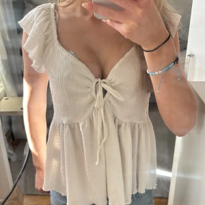 Vit volangtopp med knytning - Säljer en söt vit topp med volangärmar och knytning framtill. Toppen har v-ringning och är tillverkad i ett lätt, skrynkligt material som ger en luftig känsla.💕 Perfekt för varma dagar och enkel att styla med jeans eller kjol.Den är i storlek S och jag har bara använt den 1 gång 💕