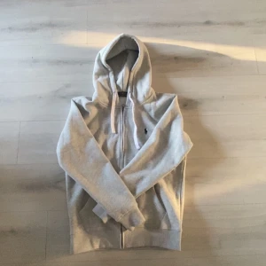 Ljusgrå hoodie med dragkedja från Ralph Lauren - Säljer en ljusgrå hoodie från Ralph Lauren med dragkedja. Perfekt för en avslappnad stil.