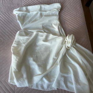 Vit axelbandslös kortklänning med rosettdetalj - Säljer en vit axelbandslös kortklänning med asymmetrisk nederdel och snygg rosettdetalj på sidan. Klänningen har en figurnära passform upptill och är perfekt för studenten🤍🤍🤍🤍🤍