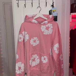 Denim tears hoodie  - Säljer en rosa hoodie från Denim Tears med vita blommor tryckta över hela tröjan. Tröjan har huva och en stor magficka framtill. Perfekt för dig som gillar färg och unika prints!