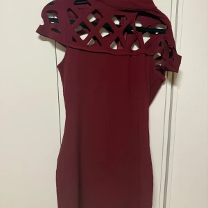 Vinröd kortklänning med cut out-detaljer - Säljer en vinröd kortklänning med unika cut out-detaljer upptill och asymmetrisk axel. Klänningen är tight och har en modern, stilren look som sticker ut. Perfekt för dig som vill ha något annorlunda och trendigt.