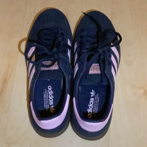 Adidas Gazelle sneakers i mörkblå och rosa - Säljer ett par Adidas Spezial sneakers i mörkblå mocka med klassiska rosa ränder och detaljer. Skorna har snörning och platt sula, perfekt för dig som gillar retrostil och vill sticka ut med färg. Ikonisk Adidas-logga på sidan och häl. Endast använda två gånger, säljer för att de var för små. Köp direkt i app. Köptes för lite mer än 900 kr!!