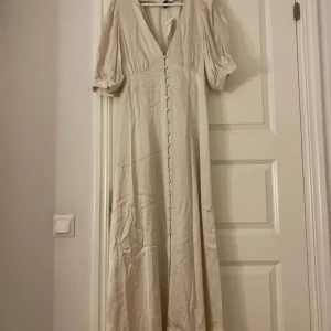 Beige långklänning med knappar - Elegant beige långklänning med v-ringning och dekorativa knappar längs hela framsidan. Klänningen har kort puffärm med knappdetalj och en mjukt fallande passform. Perfekt för somriga tillfällen och stilrena outfits.