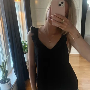 Svart linnetopp med volangdetalj - Säljer en svart linnetopp med v-ringning och volangdetaljer vid axelbanden. Toppen är ärmlös och har en enkel, stilren design som passar till mycket.