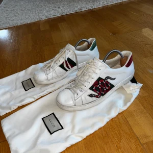 Gucci Ace sneakers med orm-motiv - Säljer ett par vita Gucci Ace sneakers med ikonisk röd och grön orm-brodyr på sidan. Skorna har gröna och röda detaljer på hälen och klassisk snörning. Kommer med original dustbags. Bra skick, det ända som har lossnat lite är de röd/gröna detaljerna bak. Därav lite lägre pris, annars fina och i bra skick!😊