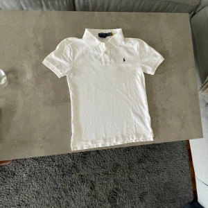 Vit piké t-shirt från Ralph Lauren L(14-16) Junior.  - Klassisk vit piké t-shirt från Ralph Lauren med korta ärmar och liten broderad logga på bröstet. Perfekt för en stilren och enkel look. Skön bomullskvalitet och normal passform.