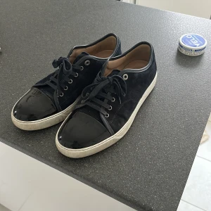 Svarta sneakers från Lanvin med lackdetaljer - Säljer ett par mörkblå sneakers från Lanvin med snygga lackade tår och vita sulor. Kommer med allt og och dustbag. Kvitto finns även. Kan gå ner i pris vid snabb affär😃