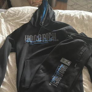 Svart hoodie från Hoodrich - Säljer en svart hoodie från Hoodrich med blå och grå detaljer och broderad logga på bröstet. Hoodien har dragsko i huvan och en mjuk insida. Perfekt för dig som gillar streetwear och vill ha en snygg och bekväm tröja.Samt ett par matchande byxor