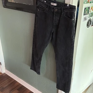 Svarta jeans från Jack & Jones - Säljer ett par svarta jeans från Jack & Jones med klassisk femficksdesign och raka ben. Jeansen har knappgylf och är tillverkade i bomullstyg med en lätt tvättad look. Perfekta för en stilren vardagsoutfit.