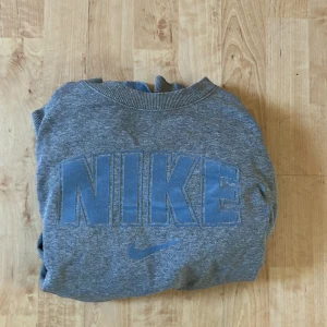 Grå sweatshirt från Nike - Säljer en grå sweatshirt från Nike med stort blått NIKE-tryck på bröstet. Tröjan har rund halsringning och långa ärmar med ribbade muddar. Perfekt för en avslappnad och sportig stil. Har endast använt tröjan ett par gånger och det finns inga fläckar eller hål