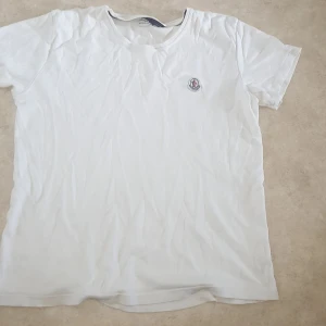 Vit t-shirt från Moncler - Säljer en klassisk vit t-shirt från Moncler med rund halsringning och Moncler-logga broderad på bröstet. T-shirten har en enkel och stilren design, perfekt för en avslappnad look.