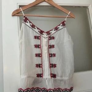 Vit broderad linnetopp sommar - Säljer en söt vit linnetopp med tunna axelband och broderade mönster i rött och svart. Toppen har knappar framtill och broderier längst halsringningen och nederkanten. Använd en gång 💕
