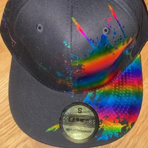 Svart keps med regnbågsfärgat stänkmönster - Säljer en svart keps från New Era med ett färgstarkt regnbågsstänkmönster över sidan och skärmen. Kepsen har platt skärm och klassisk passform. Perfekt för dig som vill sticka ut med en unik och färgglad stil.
