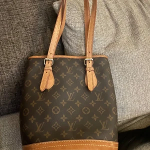 Louis Vuitton petite bucket bag  - Säljer min LV väska, mini bucket bag/petite bucket bag. Jag har ärvt den av äldre släkting och har dessvärre inga papper på den men ser man till tecken på äkthet fyller den de kriterierna. Har aldrig fått den värderad därav det låga priset på en modell som inte produceras längre. Det är svårt att se men på ett foto syns kod för vart och när väskan är gjort, S står för Frankrike precis som märket inuti säger. Runda O i Louis Vuitton. Metalltassar under väskan. Den är i mkt gott skick! 