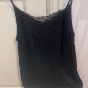 Svart linnetopp med spets - Säljer en svart linnetopp med tunna axelband och vacker spetsdetalj upptill. Perfekt att styla till olika outfits och ger en elegant touch med sitt enkla men snygga utseende.