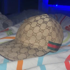 Beige keps från Gucci med GG-mönster - Säljer en beige keps från Gucci med klassiskt GG-mönster över hela kepsen och en grön-röd rand på sidan. Kepsen är i bomull och har böjd skärm. Märkt med storlek M (58 cm).