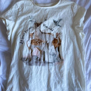 Vit t-shirt med hjortmotiv och text - Vit t-shirt i ekologisk bomull med ett gulligt motiv av hjortar och en fågel samt texten 'Let your dreams Run wild' på framsidan. T-shirten har korta ärmar och en klassisk rund halsringning.