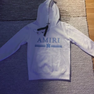 Vit hoodie från Amiri med blå logga - Säljer en vit hoodie från Amiri med blå logga och dekorativa linjer på bröstet. Tröjan har huva med dragsko och en stor magficka. Perfekt för dig som gillar streetwear och vill ha något stilrent men ändå iögonfallande.