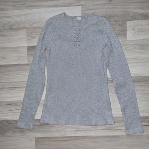 Grå ribbad långärmad tröja från Lindex - Säljer en grå ribbad långärmad tröja från Lindex med snörningsdetalj vid halsen. Tröjan har en rund halsringning och är i mjukt, stretchigt material. Perfekt för en enkel och stilren look.