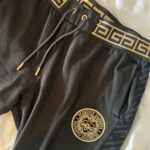 Svarta mjukisbyxor från Glorious Gangsta - Säljer ett par svarta mjukisbyxor från Glorious Gangsta med guldfärgade detaljer och snörning i midjan. Byxorna har ett snyggt märke på benet och ett brett resårband med guldmönster upptill. Perfekta för en trendig och avslappnad stil.