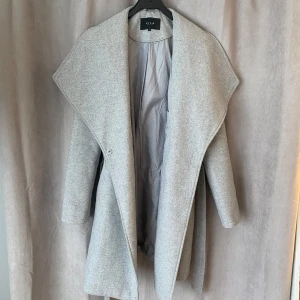 Ljusgrå kappa från Vila - Snygg ljusgrå kappa från Vila med bred krage och bälte i midjan. Kappan har lång ärm och är tillverkad i ett mjukt ullblandat material. Perfekt för kyliga dagar och ger en stilren look. Finns en liten fläck på kragen(går nog lätt att tvätta bort)