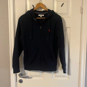 Marinblå hoodie från U.S. POLO ASSN - Marinblå hoodie med dragkedja från U.S. POLO ASSN. Klassisk design med huva och två fickor framtill. Liten röd broderad logga på bröstet. Perfekt för en avslappnad och stilren look.