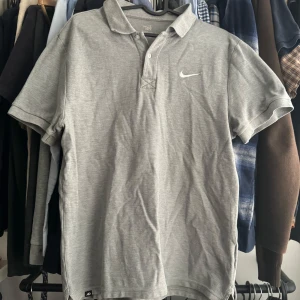 Grå pikétröja från Nike - Säljer en klassisk grå pikétröja från Nike med vit broderad logga på bröstet. Tröjan har korta ärmar, krage och knäppning framtill. Perfekt för en sportig och avslappnad stil.
