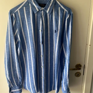Blåvit randig skjorta från Polo Ralph Lauren - Säljer en blå och vit randig skjorta från Polo Ralph Lauren med klassisk krage och broderad logga på bröstet. Skjortan har lång ärm och knäppning framtill. Perfekt för en stilren och avslappnad look.